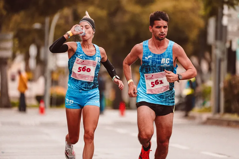 Dois atletas correndo uma meia maratona enquanto uma corredora bebe água